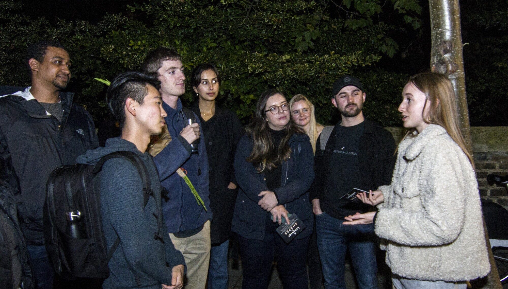 A female scientist in beige speaks to a group of people outside in the dark