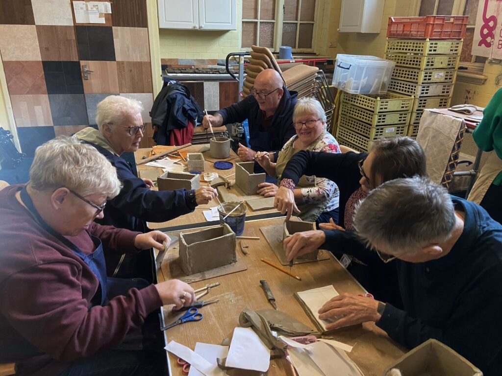 A group of older people sitting at a table, taking part in a ceramics workshop. 